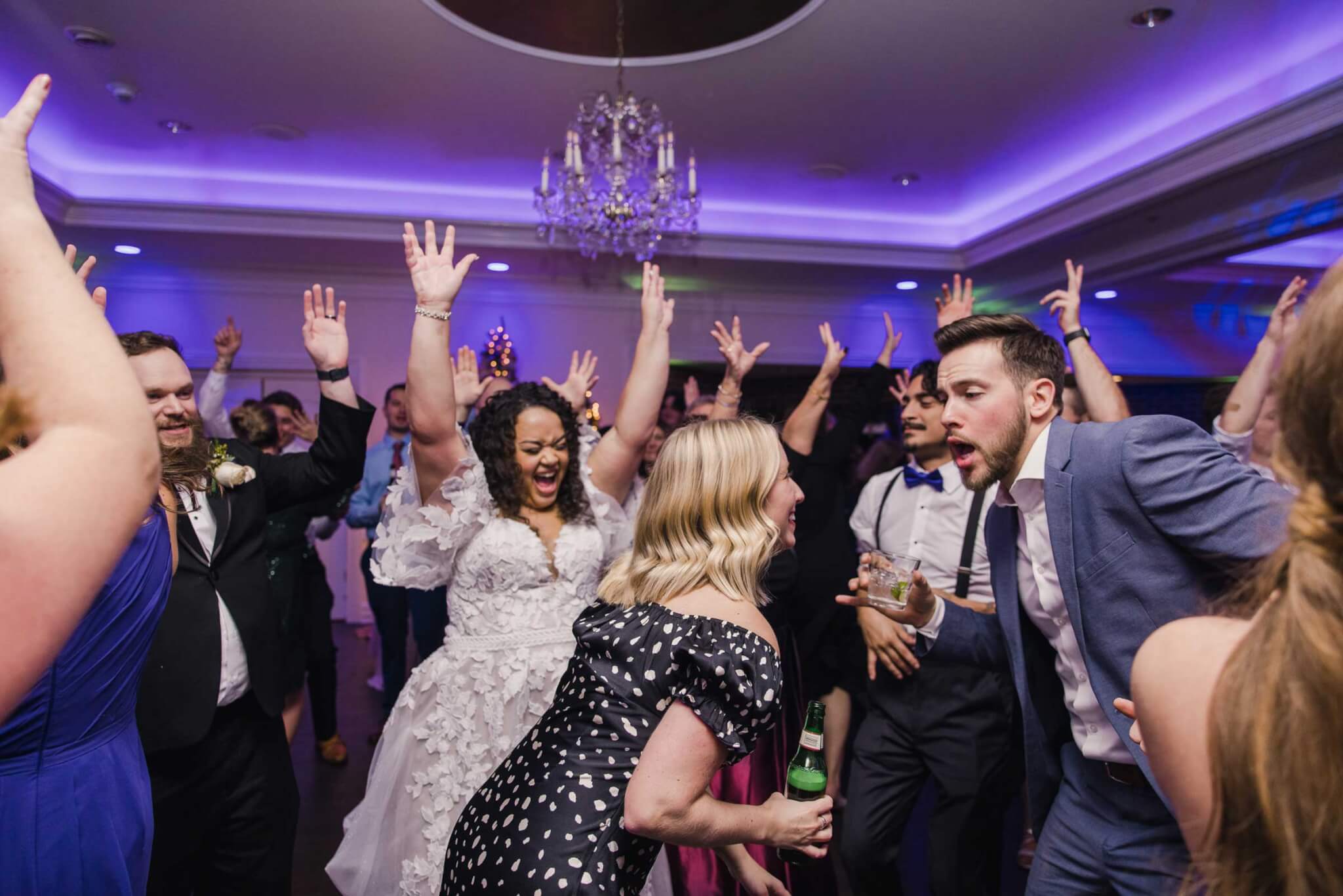 sonic-home-wedding-dancing-hero A group of people, including a bride in a white dress, dance energetically with raised hands at an indoor wedding reception under purple lights.