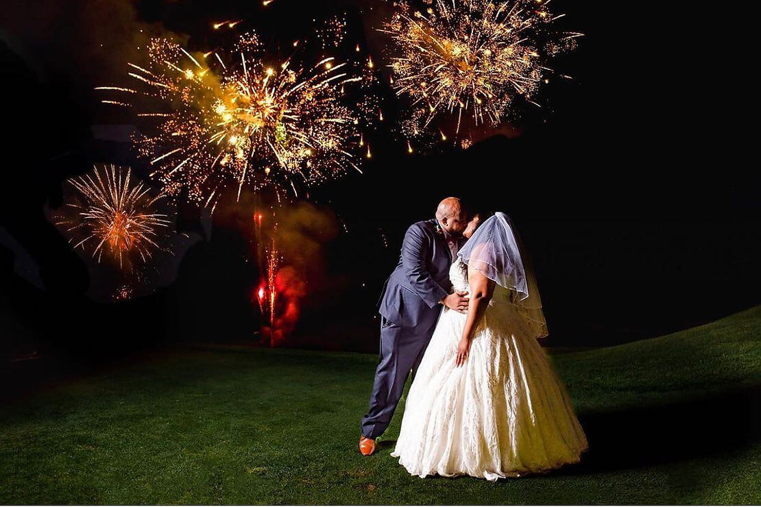 A bride and groom kiss on a grassy lawn at night with bright fireworks lighting up the dark sky behind them.