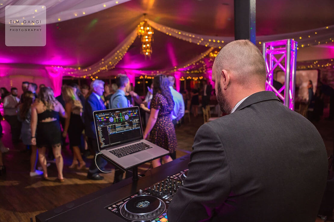 c4fb96_ae638186c2be482887f274856f11016e~mv2 A DJ in a suit plays music on a mixing console at an indoor event with people dancing under string lights and purple lighting.