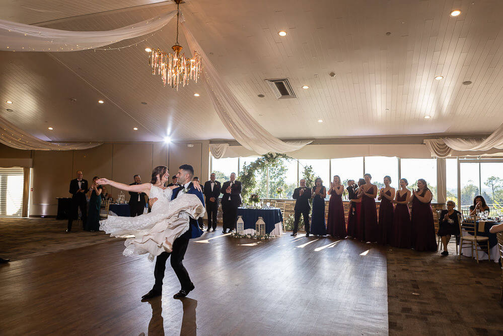 A groom lifts and twirls the bride on the dance floor while guests watch in a decorated, well-lit banquet hall.