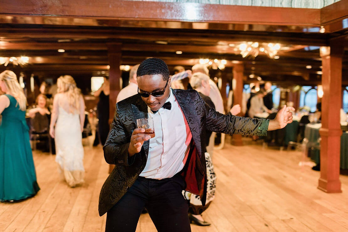 A man in a suit and sunglasses dances energetically with a drink in hand at an indoor event, while other guests are seen in the background.