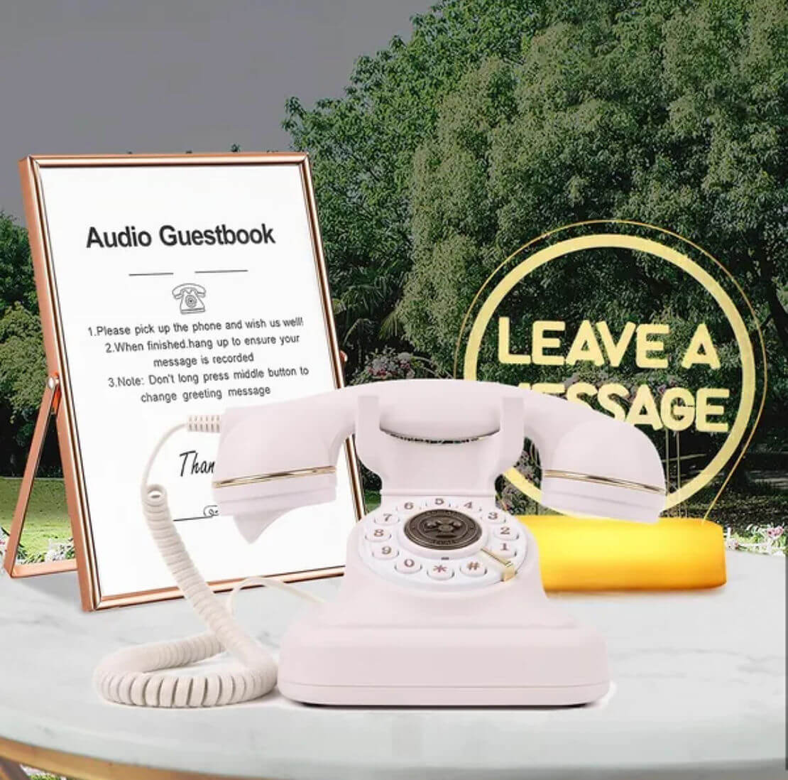 A white vintage rotary phone on a table with a sign reading "Audio Guestbook" and another sign that says "Leave a Message" against a green outdoor backdrop.