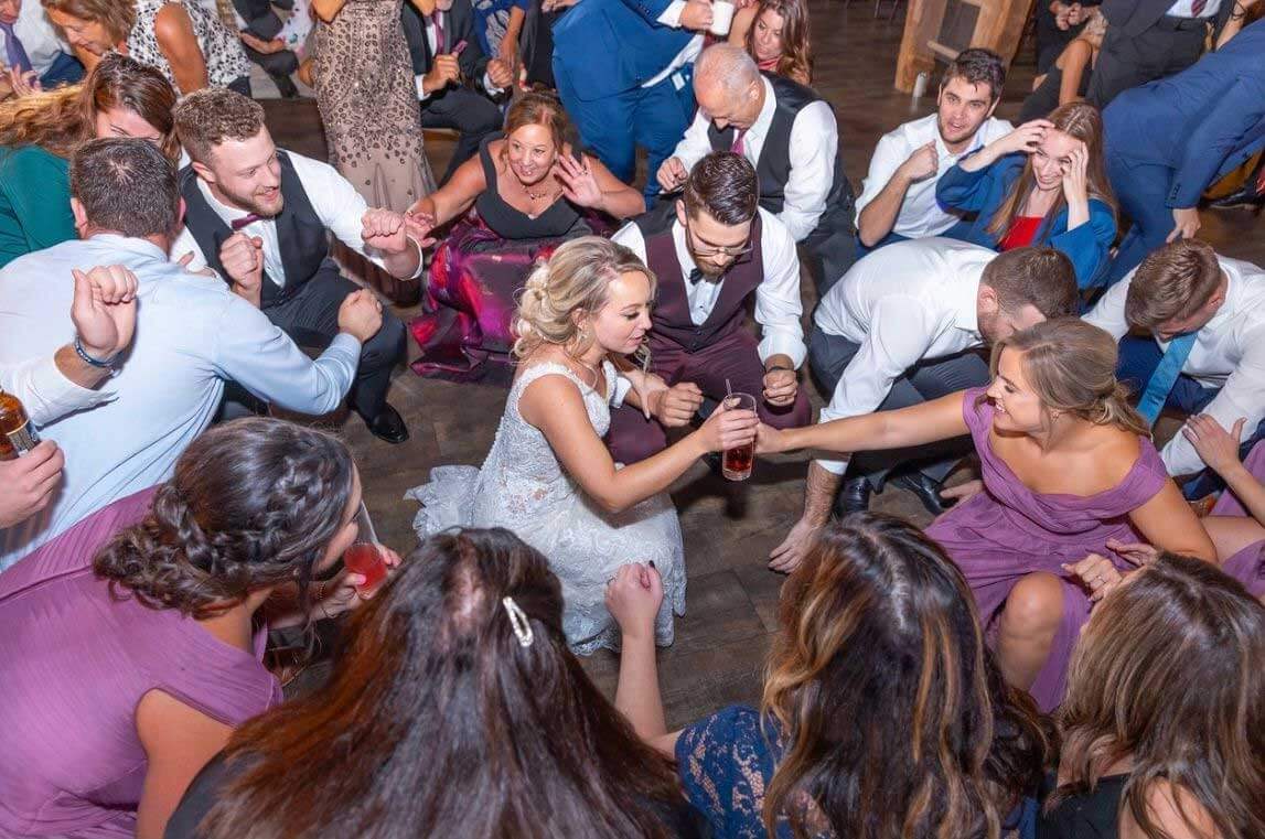 bride-having-fun-on-the-dance-floor A bride and groom crouch and hold drinks, surrounded by laughing and dancing wedding guests on a crowded dance floor.