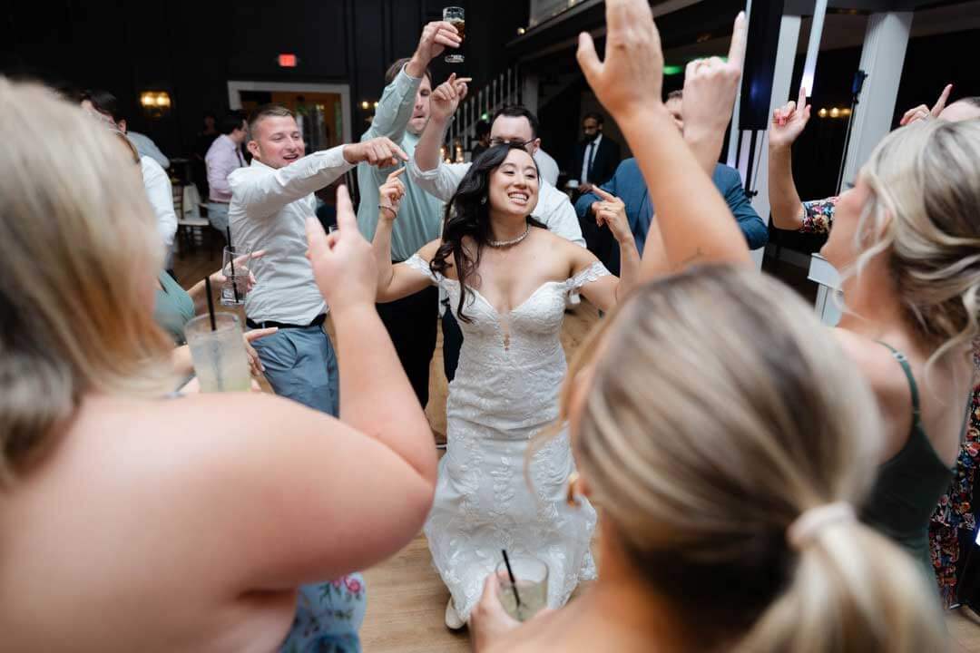 A bride in a white dress dances joyfully in the center of a group of wedding guests who are cheering and raising their arms.