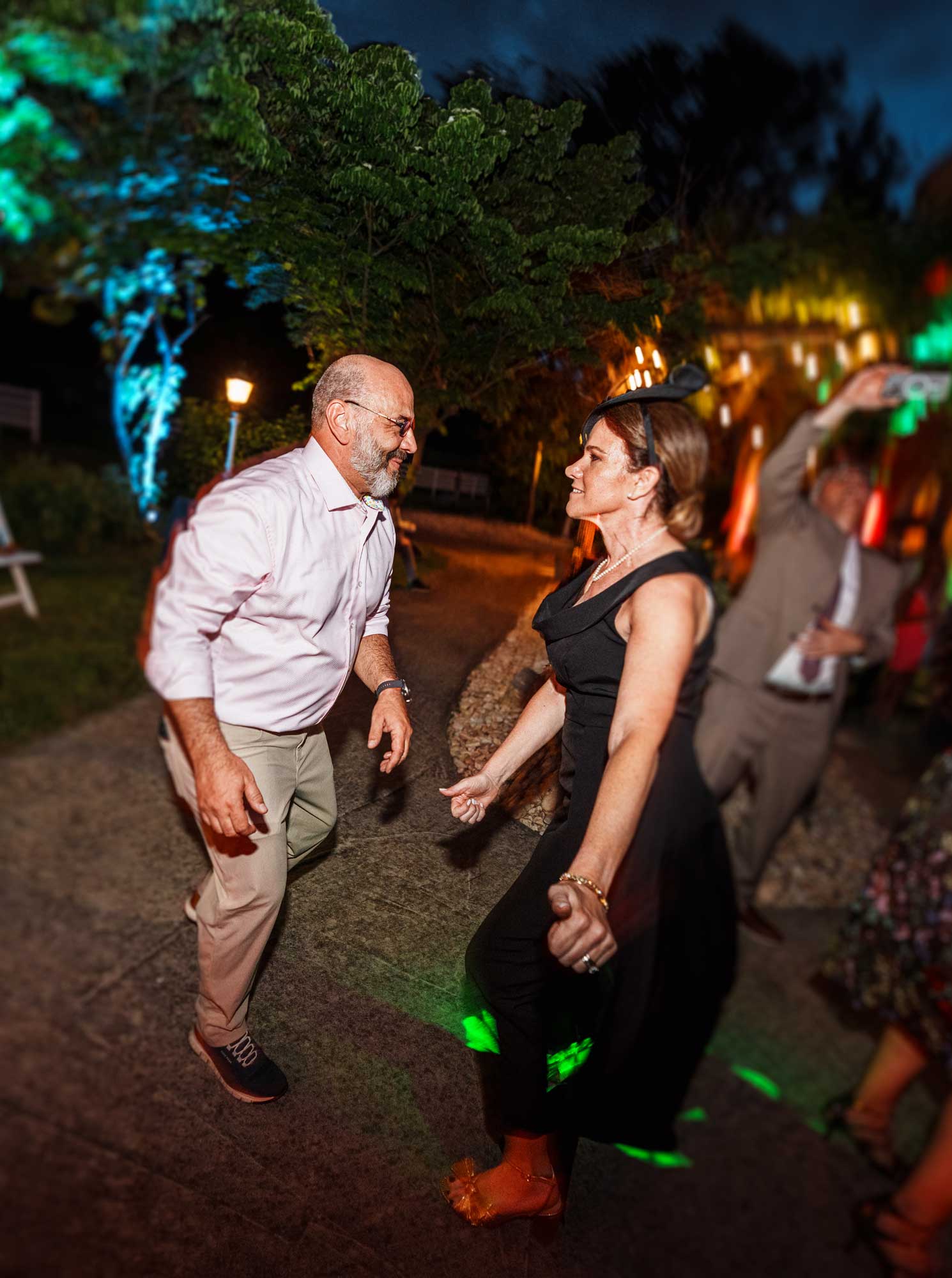 A man in a light shirt and khaki pants and a woman in a black dress are dancing outdoors at night, surrounded by colorful lights and other people.