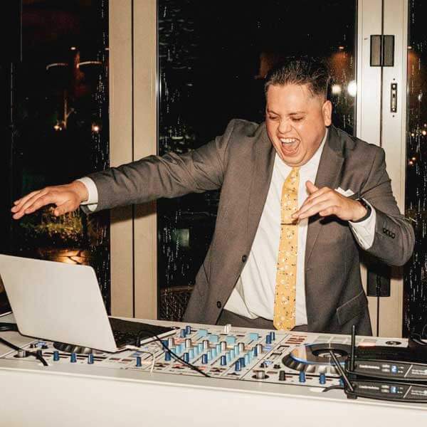 A man in a suit enthusiastically operates a DJ controller and laptop at an indoor event, smiling with one hand raised.