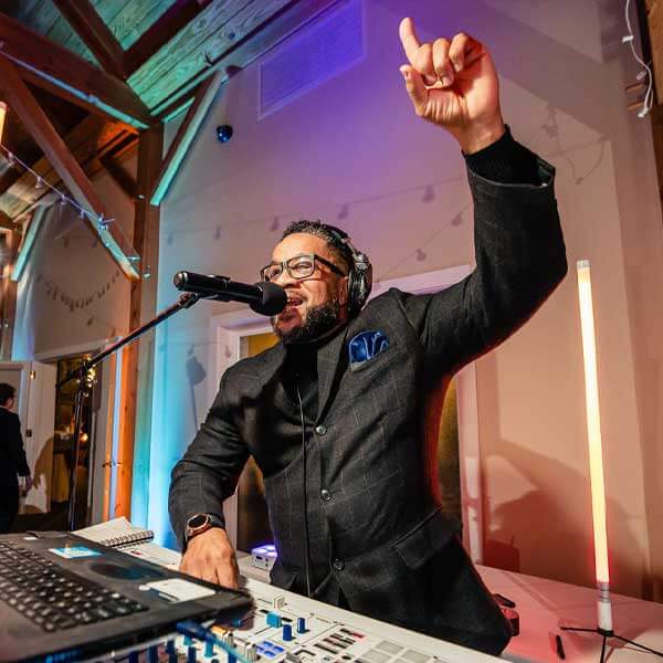 A DJ wearing glasses and a dark suit speaks into a microphone, raising one hand while standing behind a DJ mixer and laptop at an indoor event.