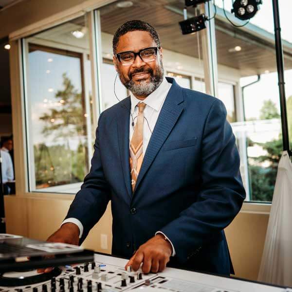 A man in a blue suit and tie stands indoors, smiling while operating DJ equipment. Large windows and lighting equipment are visible in the background.