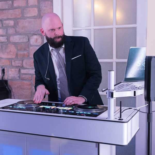 A man in a suit operates a DJ controller at an indoor event, with a laptop and sound equipment set up in front of him.