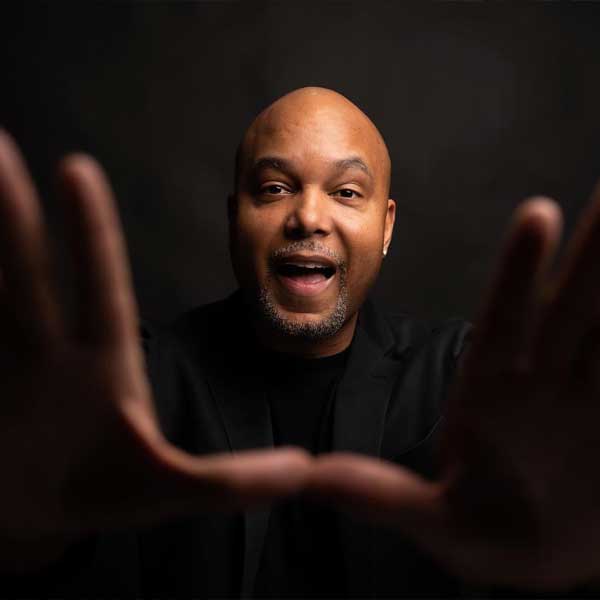 A man in a black shirt faces the camera, smiling with mouth open and holding his hands up in front, palms facing outward. The background is dark.