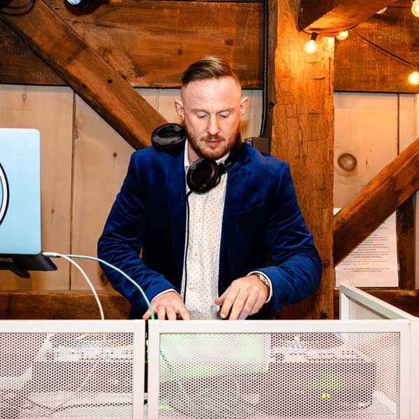 A man wearing a blue blazer and headphones stands at a DJ booth, operating equipment in a rustic wooden setting.