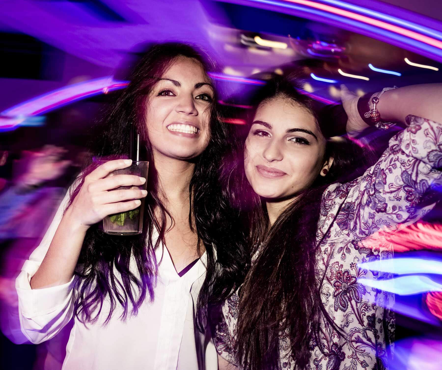 Two young women pose and smile in a lively, colorful club setting; one holds a drink, and blurred lights create a dynamic background.
