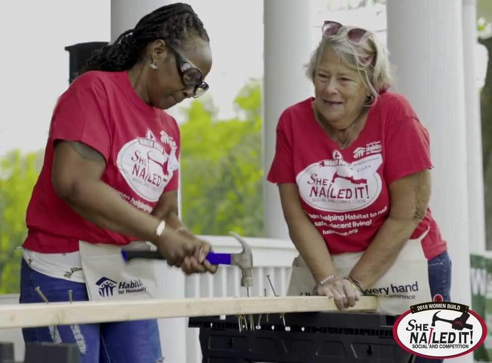 Two women in red "She Nailed It!" shirts work together to hammer nails into a plank of wood during a Habitat for Humanity event.