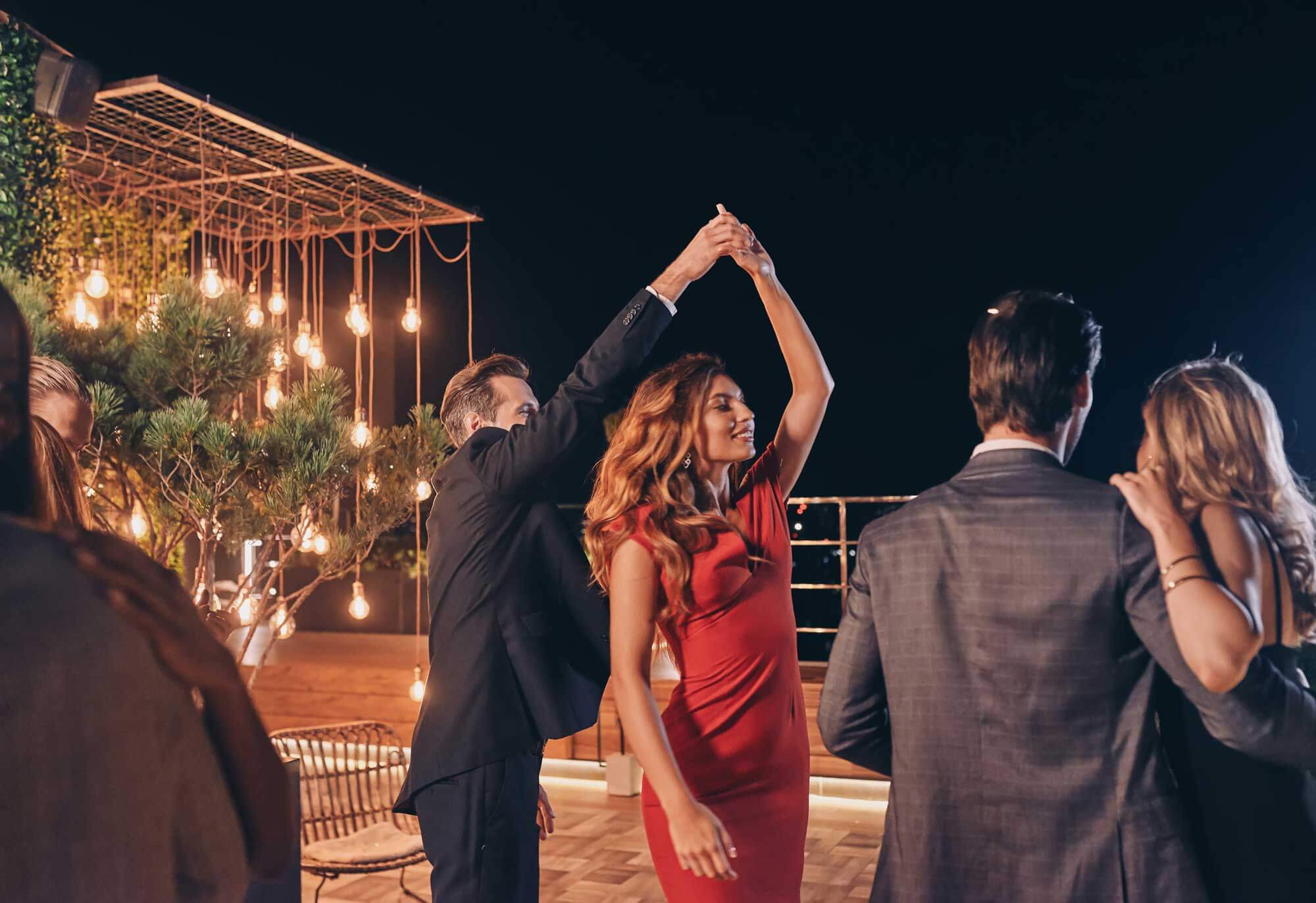 People in formal attire are dancing on a rooftop at night, with string lights and trees in the background.