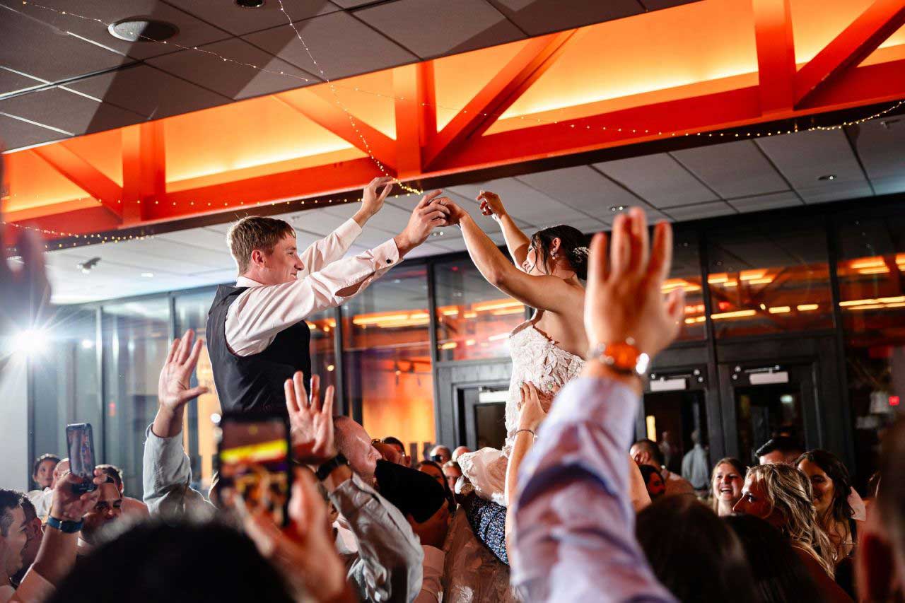 Bride and groom are lifted on chairs during a wedding celebration, surrounded by cheering guests inside a brightly lit venue with orange accents.