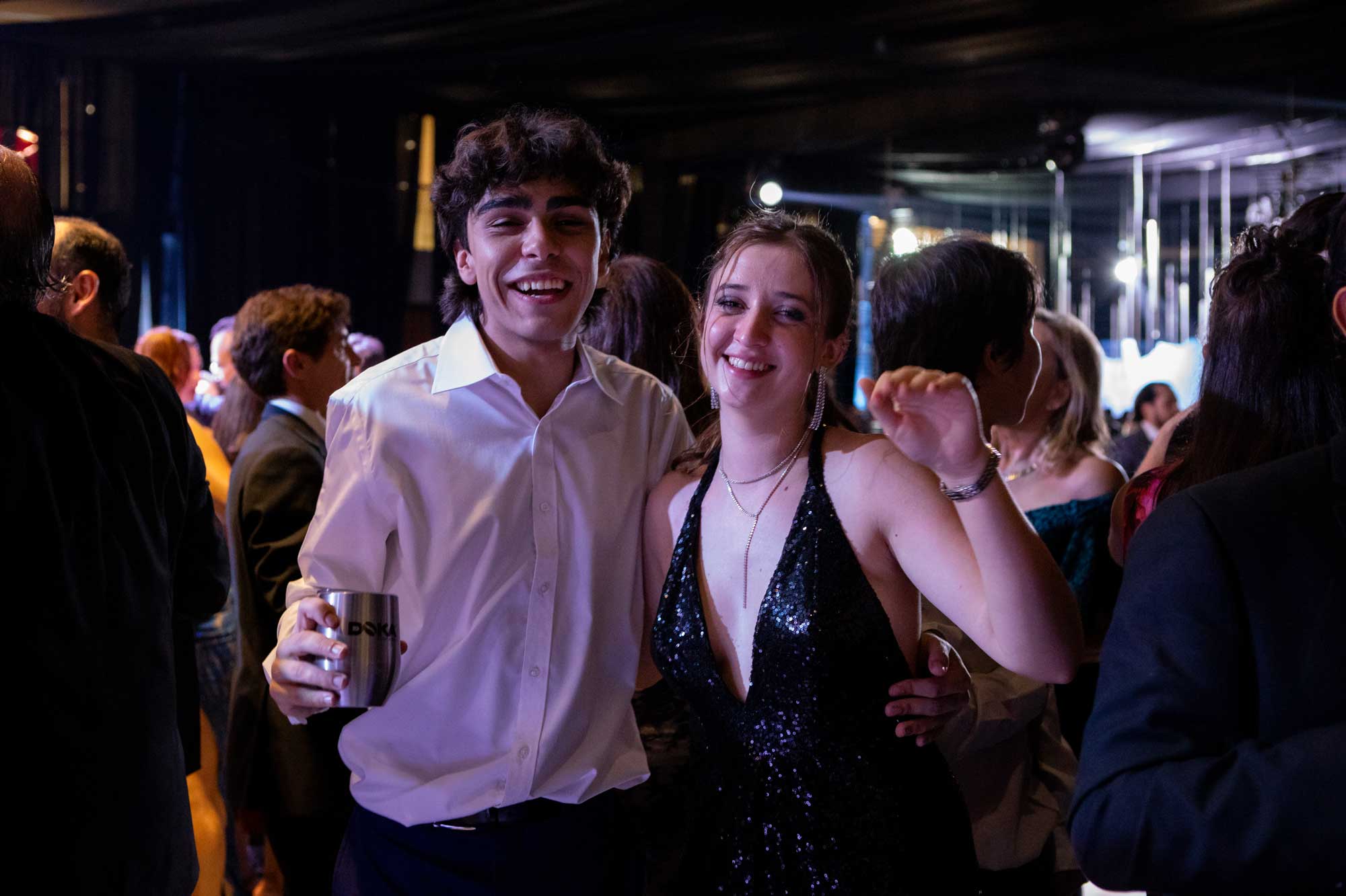 Two people pose and smile at a crowded indoor event; one holds a drink, and both wear formal attire under dim lighting.