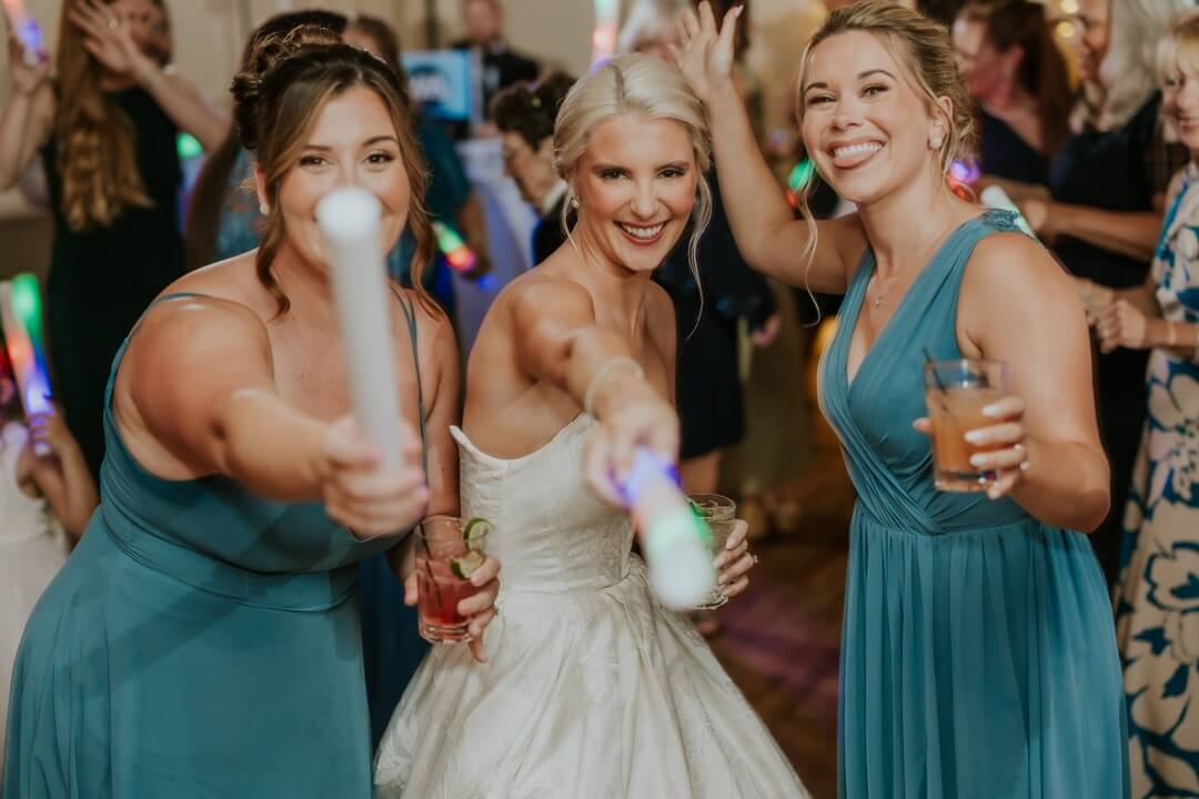 Contact Lehigh Valley DJ Sonic Entertainment Three women, including a bride in a white dress and two bridesmaids in blue dresses, smile and hold glowing sticks at a lively indoor event.