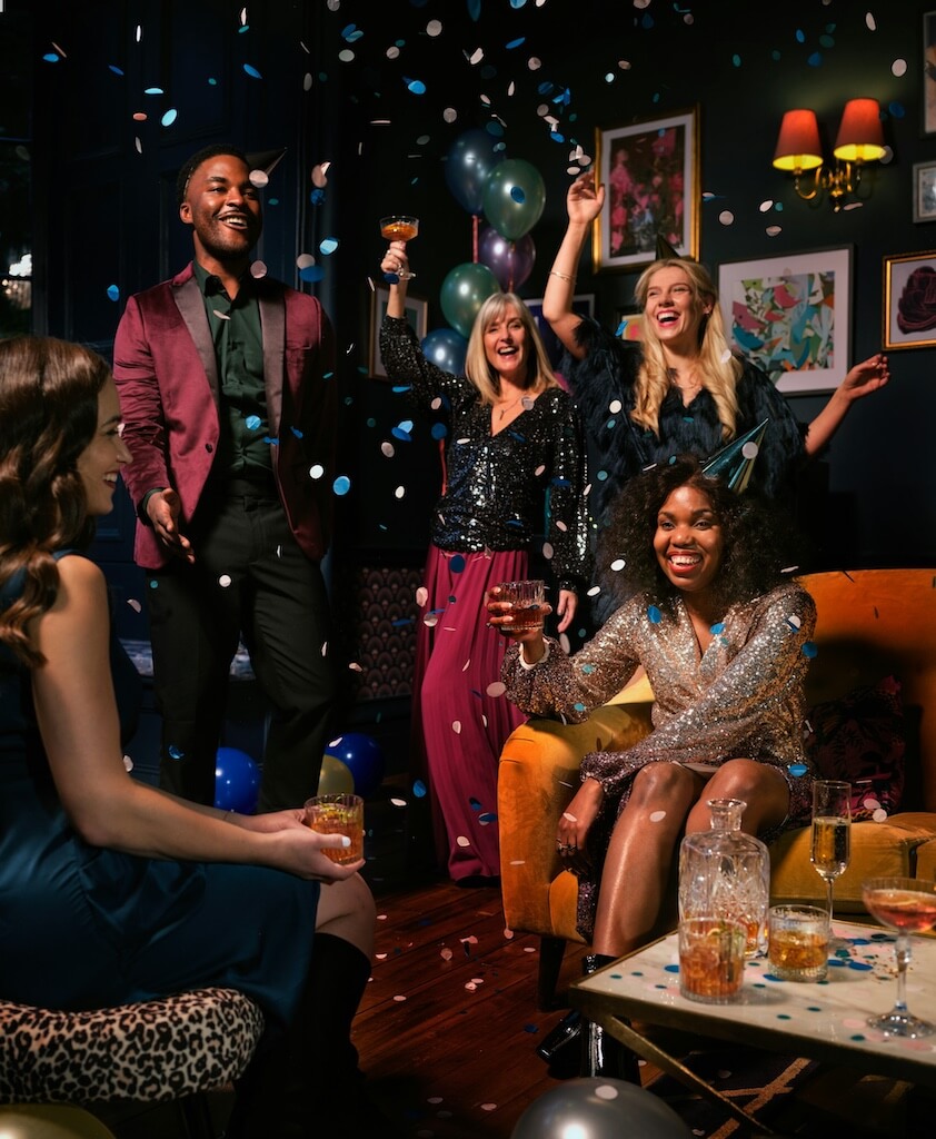 Frequently-Asked-Party-DJ-Questions Five people dressed in party attire celebrate indoors with drinks, confetti, and balloons. Decorations and drinks are visible on a table in the foreground.
