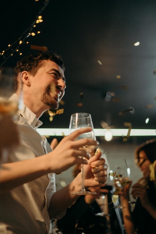 Frequently-asked-Lehigh-Valley-DJs-questions A man holding a glass and smiling at a party, surrounded by people and confetti in the air.