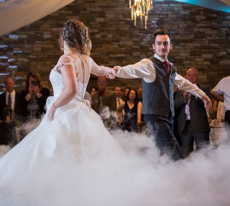 A bride and groom dance together on a fog-covered floor at their wedding reception, with guests watching in the background.