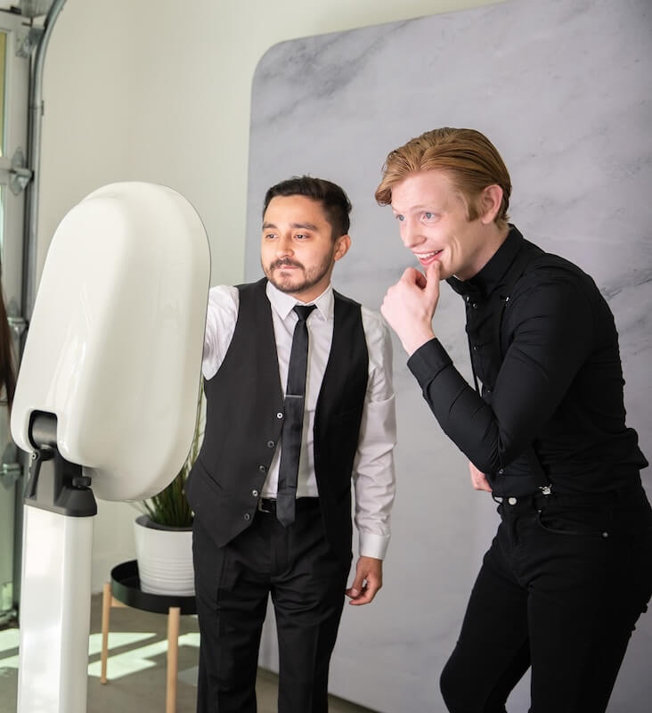 Two men in formal attire stand in front of a photo booth, one pointing at the screen while the other poses and smiles.
