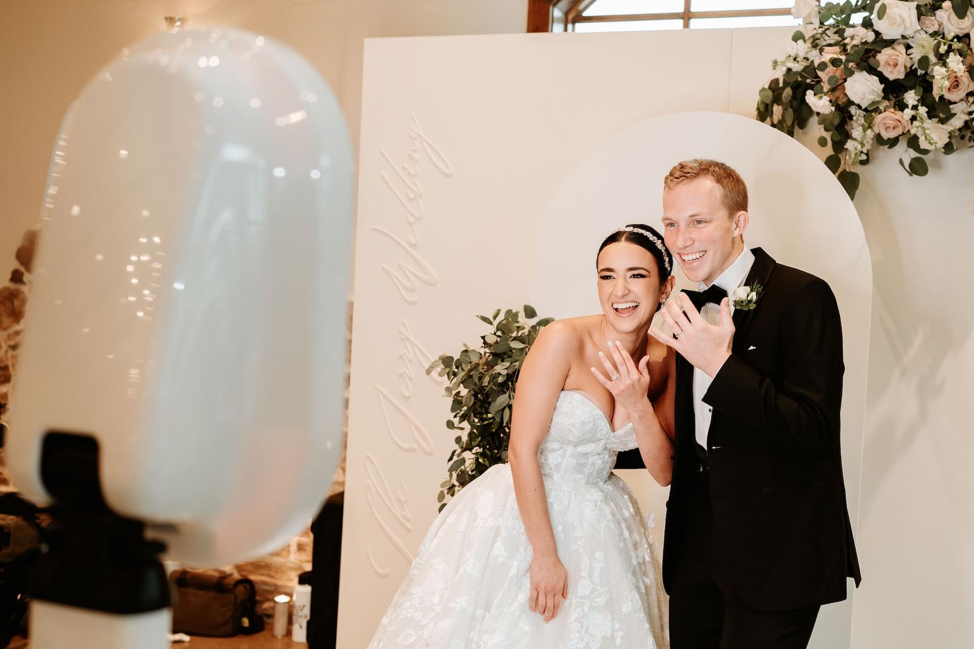 A bride and groom in formal attire smile and wave at a photo booth during their wedding, with floral decorations in the background.