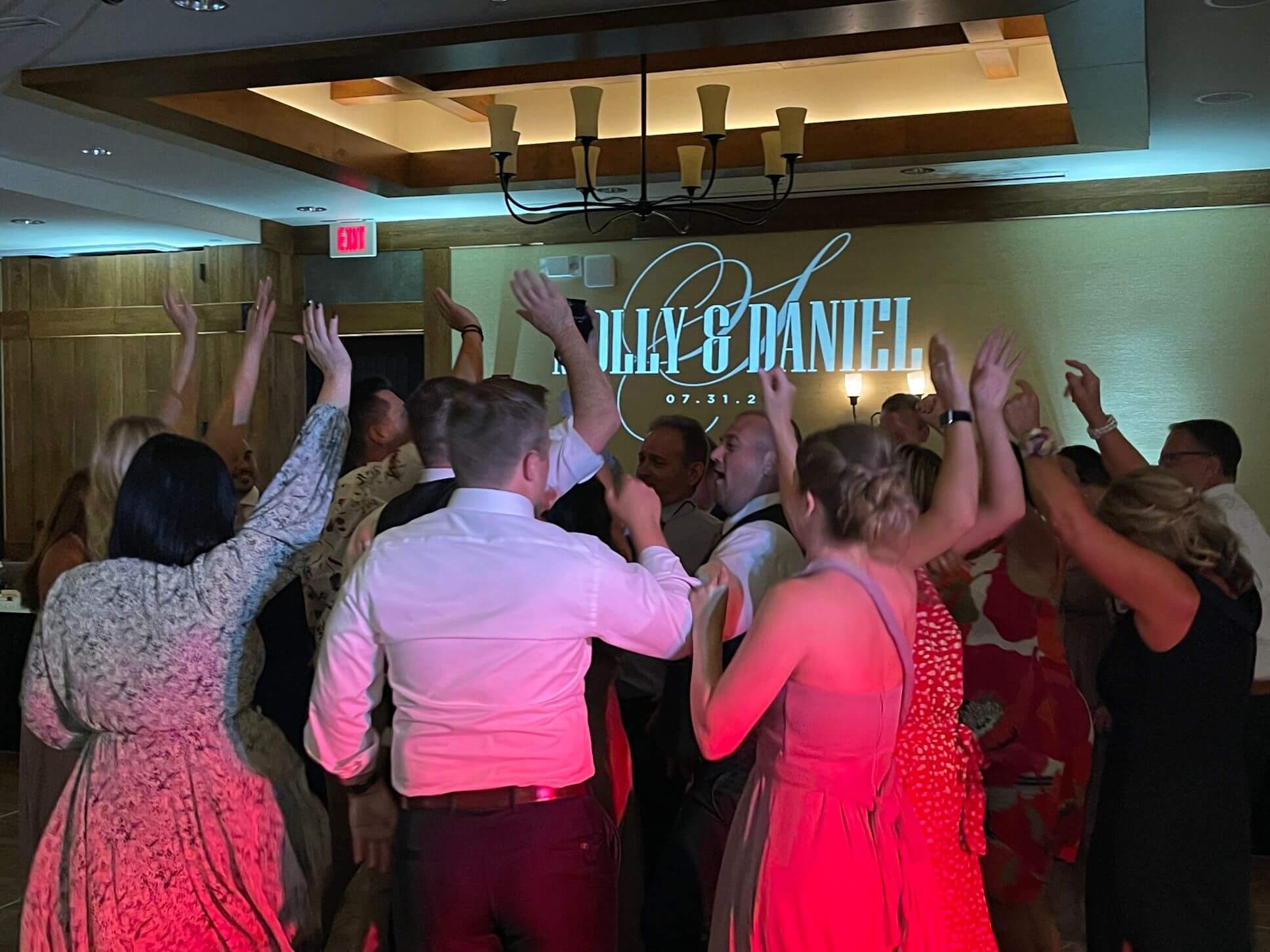 A group of people dance with raised hands at a wedding reception. The wall projection behind them displays the names "Kayla & Daniel" and the date "07.31.21.