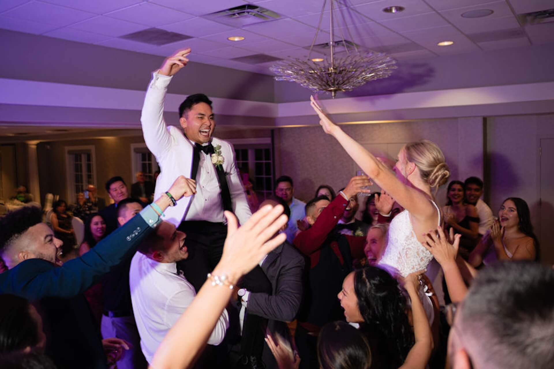 A groom is lifted on chairs by guests while the bride and others cheer around him at a wedding reception indoors.