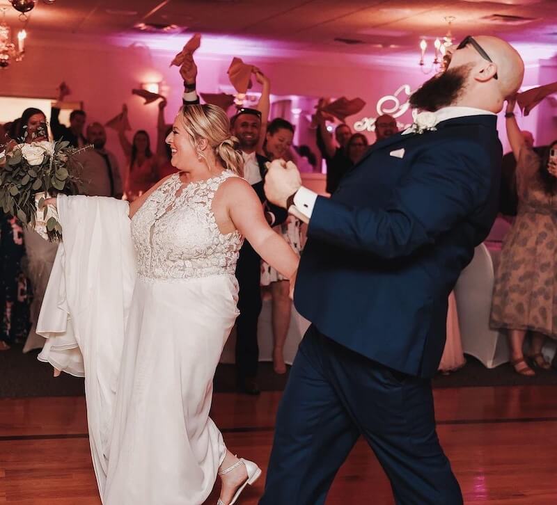 A bride and groom celebrate while entering a wedding reception, surrounded by cheering guests waving napkins in the air.