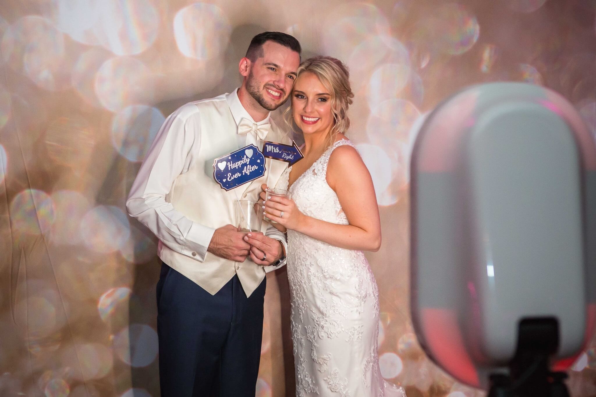 A bride and groom pose for a photo together, holding props and smiling in front of a blurred, sparkling backdrop.