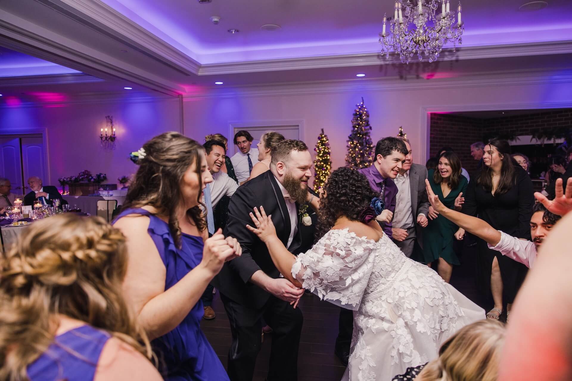 A bride and groom dance surrounded by guests at a wedding reception in a decorated hall with a Christmas tree in the background.