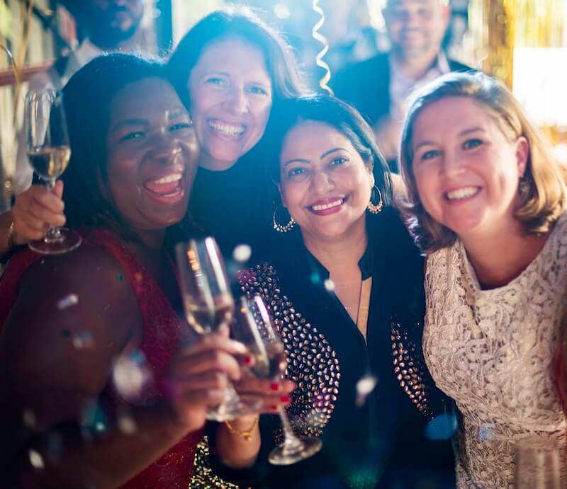 Restaurant Chilling Out Classy Lifestyle Reserved Concept Four women smiling and holding glasses of champagne at a festive indoor gathering, with people and decorations visible in the background.
