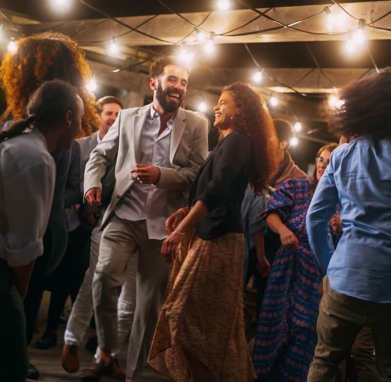 Beautiful Carefree Friends are Dancing Together and Celebrating an Evening Event at a Party . Diverse Multiethnic Young Adult People Have Fun at a Corporate Party in a Restaurant. A group of people are dancing and socializing under string lights at an outdoor event in the evening.