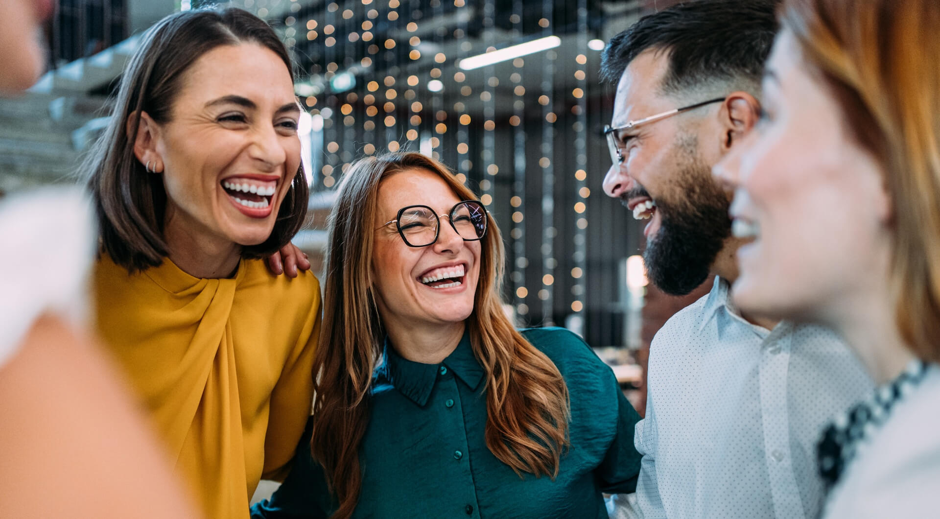 Excited coworkers embracing each other during a celebration in the office. Four adults standing close together, smiling and laughing in a lively indoor setting with string lights in the background.