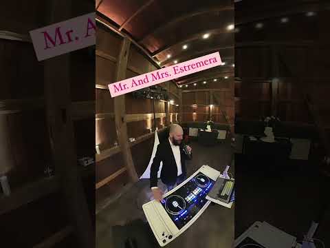 A DJ stands behind a white booth with turntables and a microphone in a dimly lit wooden venue; text reads “Mr. And Mrs. Estremera.”.