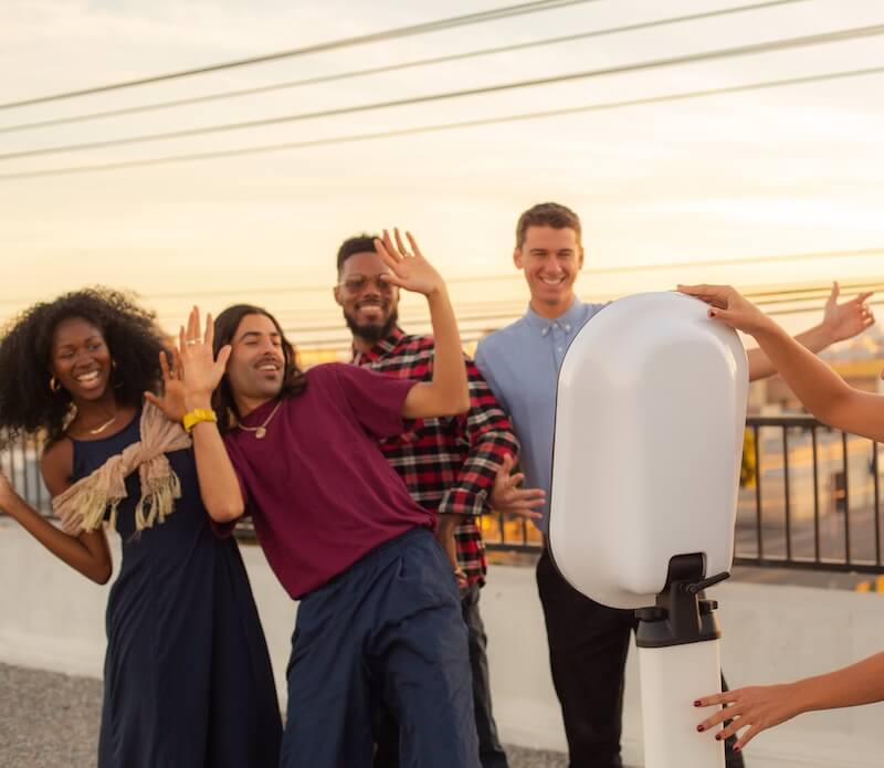 photo booth rental Four people pose cheerfully in front of a camera on a rooftop, with one person operating the camera and the sunset in the background.