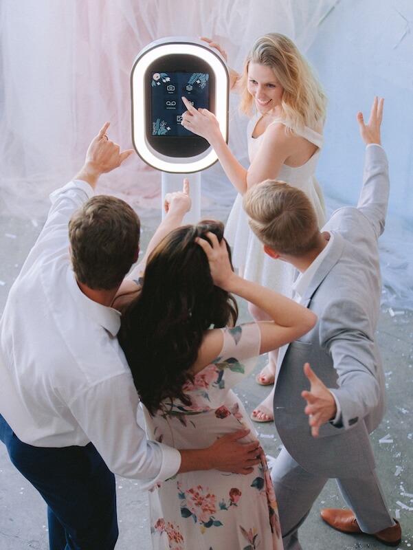 Four people stand around a photo booth, posing and pointing at the touchscreen while one woman in a white dress selects options.