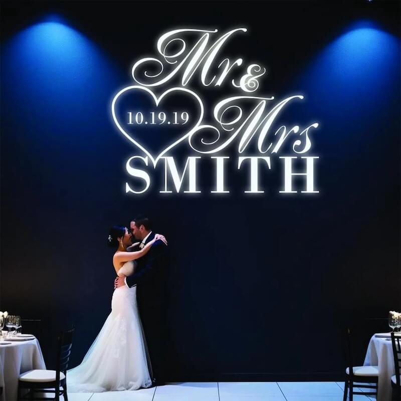wedding monogram A bride and groom embrace on a dance floor beneath a sign reading "Mr & Mrs Smith 10.19.19" in white neon letters against a dark wall.