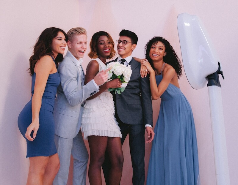 wedding selfie booth Five people dressed in formal attire pose and smile together in front of a photo booth; one person holds a bouquet of white flowers.