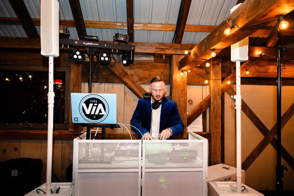 A DJ wearing headphones and a blue velvet jacket mixes music at a white DJ booth inside a rustic wooden venue with string lights overhead.